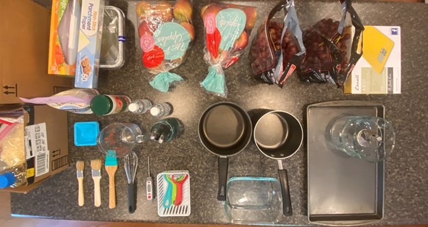 Image description: A picture containing supplies for making biopolymers spread out on a counter. There is a box of ziploc bags, parchment paper, two bags of apples, two bags of grapes, a food scale, a box containing vegetable oil and beeswax, a bag of anthem gum, a bottle of citric acid two paint brushes, one silicone pastry brush, a whisk, weigh trays, a measuring cup and tablespoons, a temperature probe, three peelers, two pots, small  glassware , a mixer and a sheet pan.